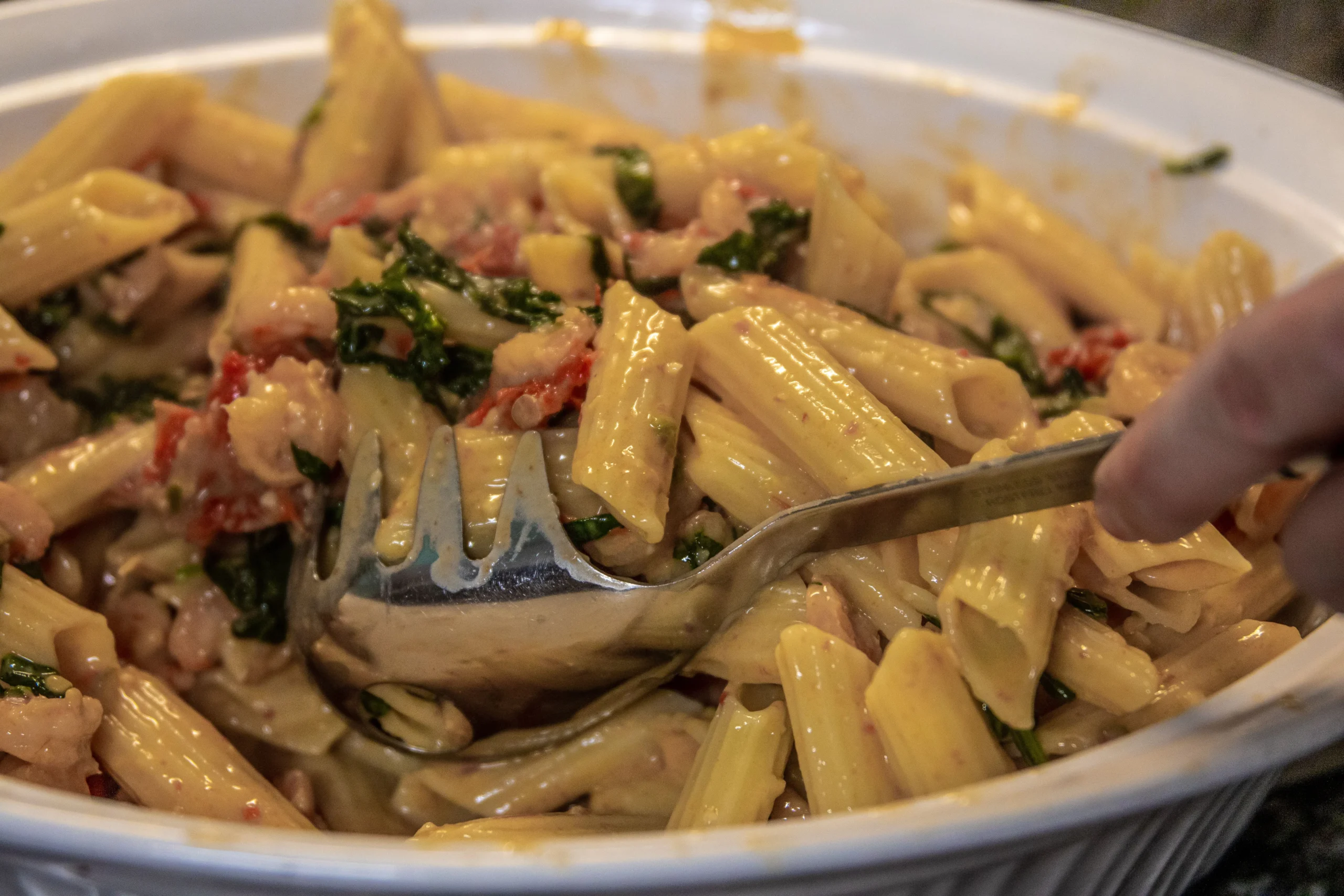 A bowl of creamy penne pasta salad with spinach and sun-dried tomatoes is served with a metal spoon..